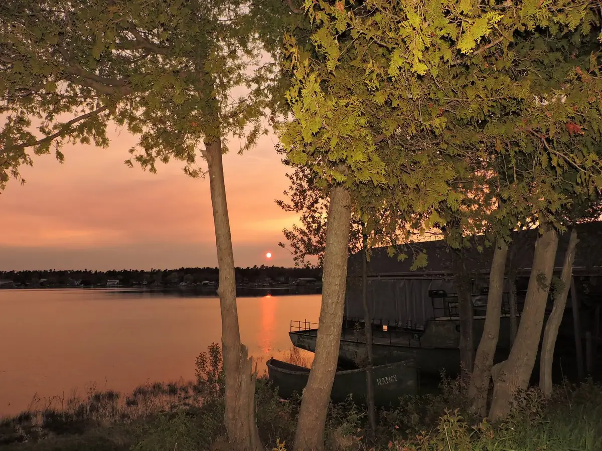 Sunset over calm water, framed by trees, with a small dark boat marked “NANCY” at the shore.