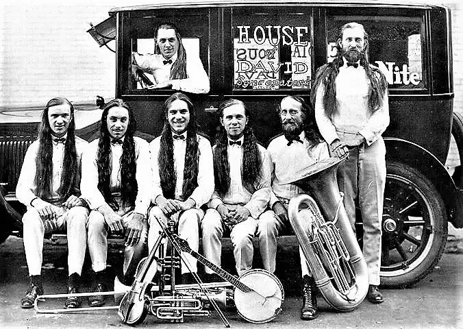 Six long-haired men pose with musical instruments beside a vintage automobile marked “HOUSE of DAVID Syncopators.”