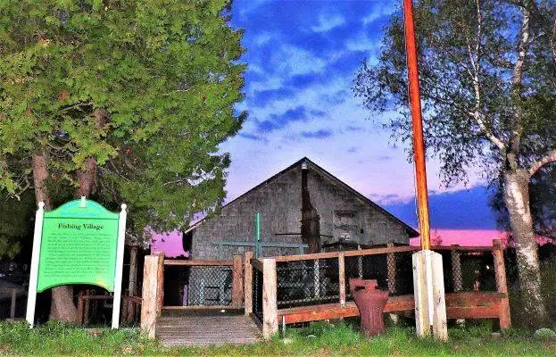 A weathered wooden building stands behind a sign reading “Fishing Village,” with trees and a vivid evening sky.