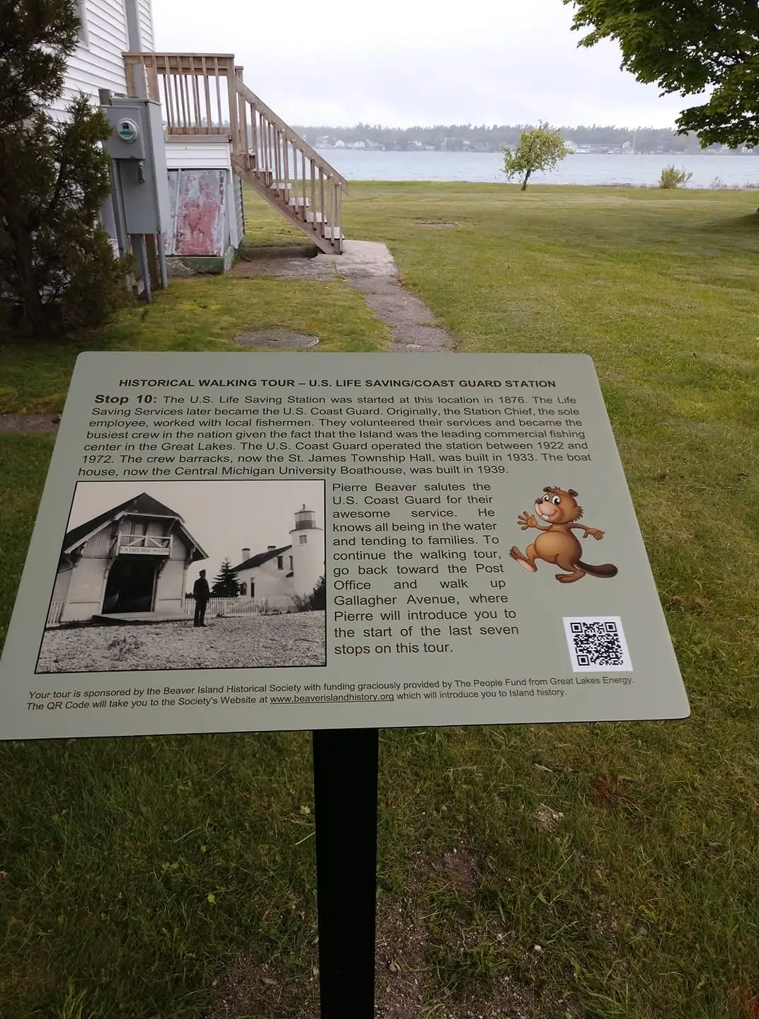 An interpretive walking-tour sign stands on a lawn beside a white-sided building, with water and a distant shoreline behind it.
