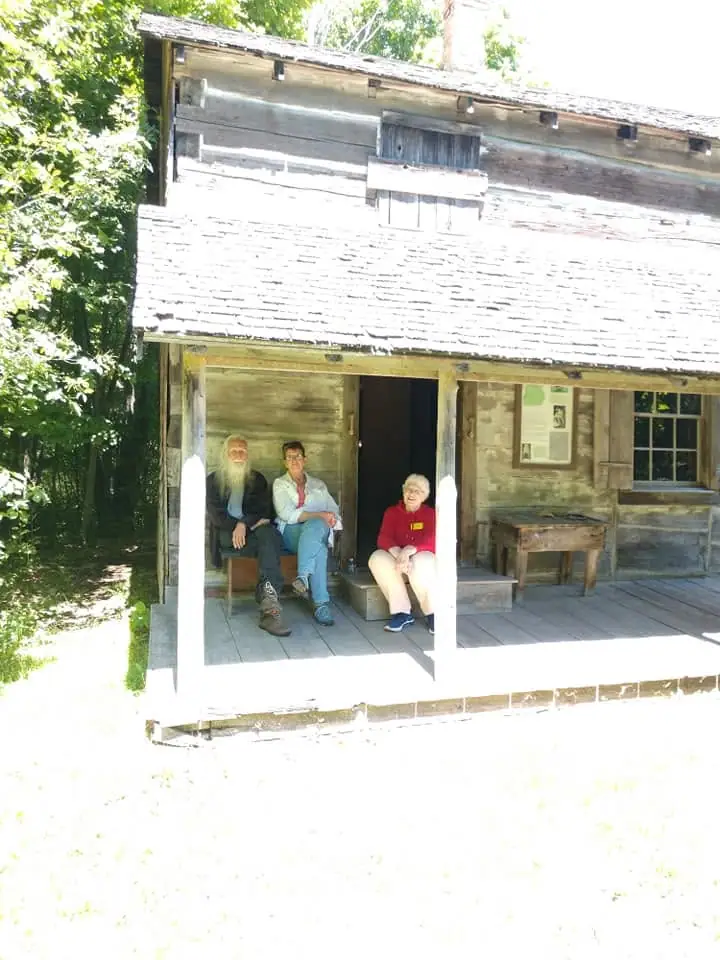 Three people sit on the porch of a weathered log building surrounded by trees.