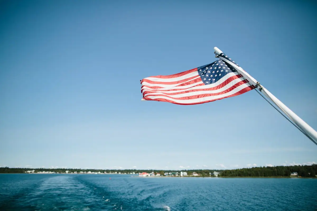 An American flag flies from a white boat mast above open water and a distant tree-lined shoreline.