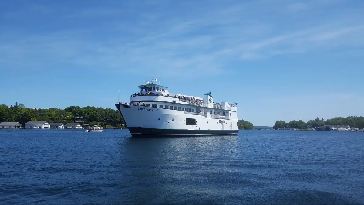 White passenger ferry EMERALD ISLE travels across blue water beside a wooded shoreline.