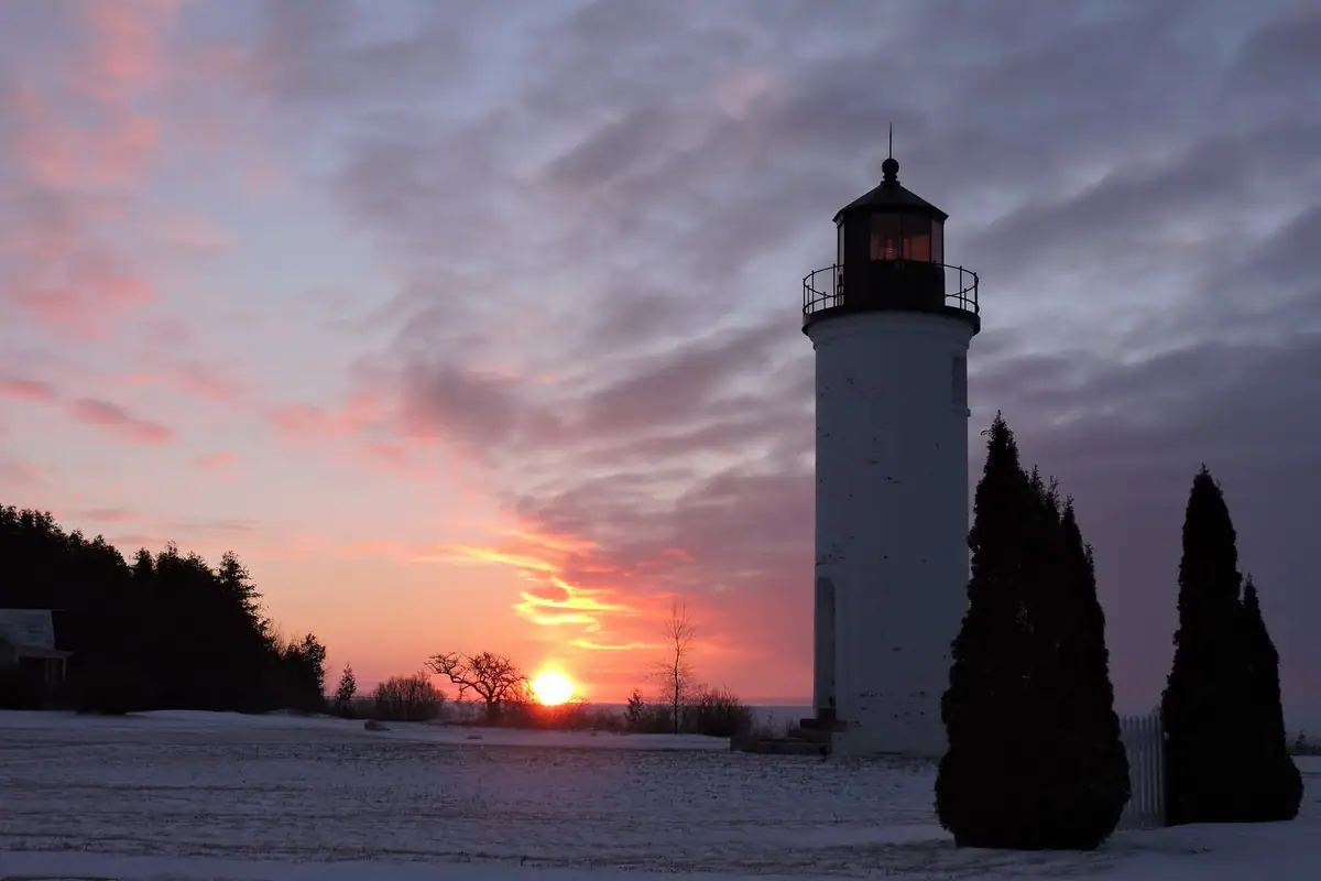 A white lighthouse stands in a snowy field beside dark evergreen trees at sunset.