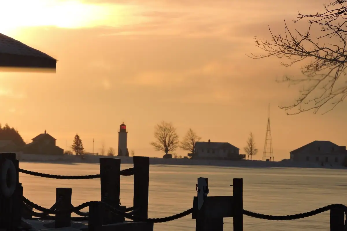 A lighthouse and buildings stand beyond frozen water at sunset, viewed past a roped dock.