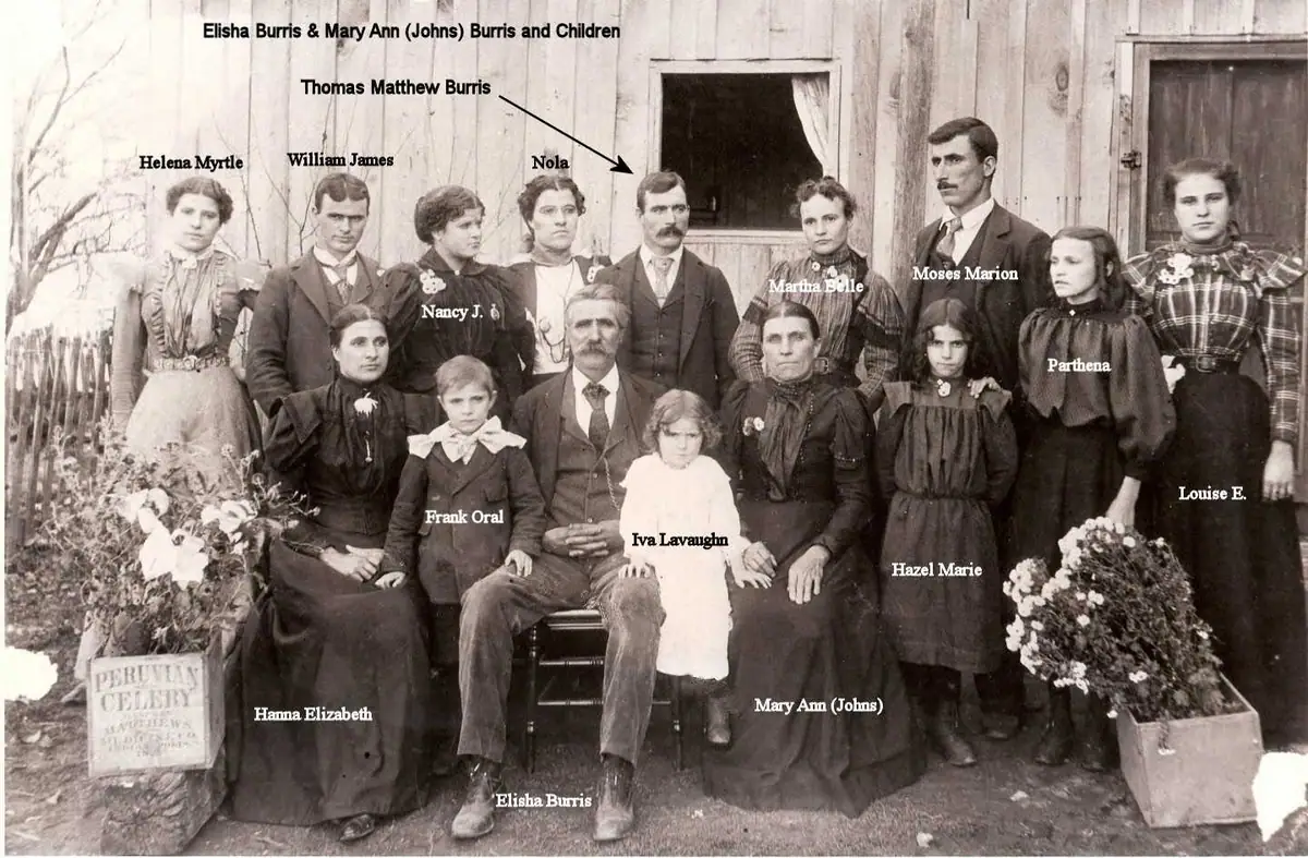 Large formally posed group portrait of adults and children outside a wooden building, with flowers in the foreground.