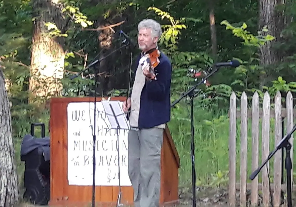 A gray-haired man plays a violin at an outdoor stage beside microphones and a sign.