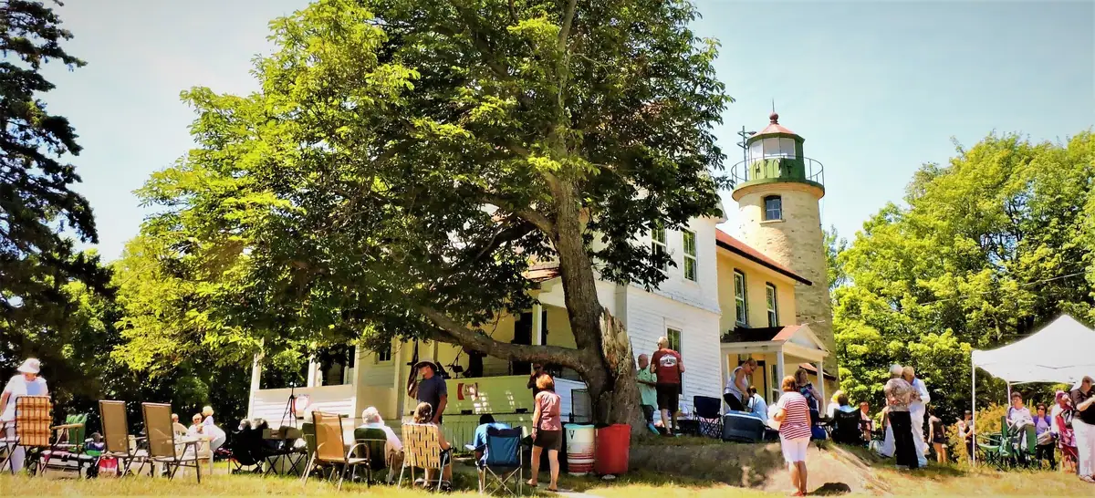 People gather beneath a large tree beside a white lighthouse and a canopy tent.