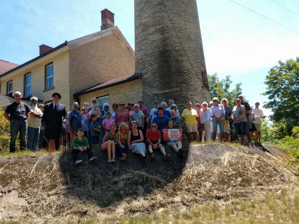 A large group poses beside a brick building and cylindrical stone tower, with two people holding “THIS PLACE MATTERS” signs.
