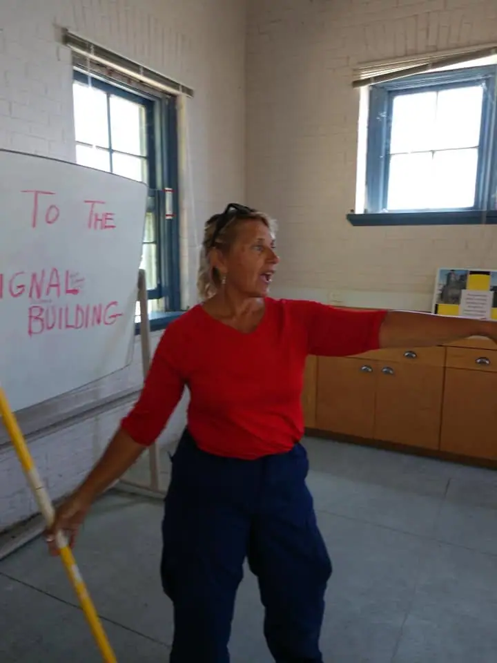 A woman presents beside a handwritten whiteboard in a bright room.