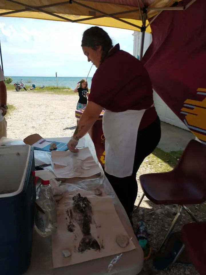 A woman works at a table beneath a canopy beside the shoreline, with a child and outdoor equipment nearby.