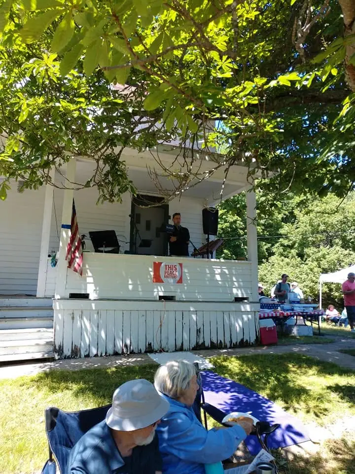 A speaker addresses an outdoor gathering from a porch beneath trees, beside an American flag and a “THIS PLACE MATTERS” sign.