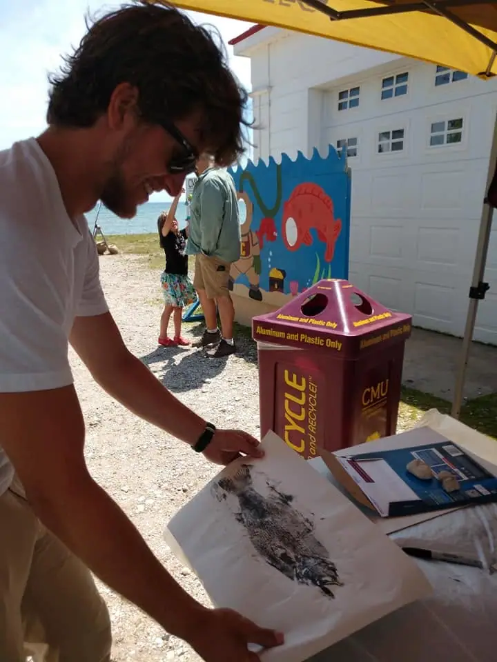 A man examines a fish-shaped impression on paper at an outdoor display table beside a recycling bin.