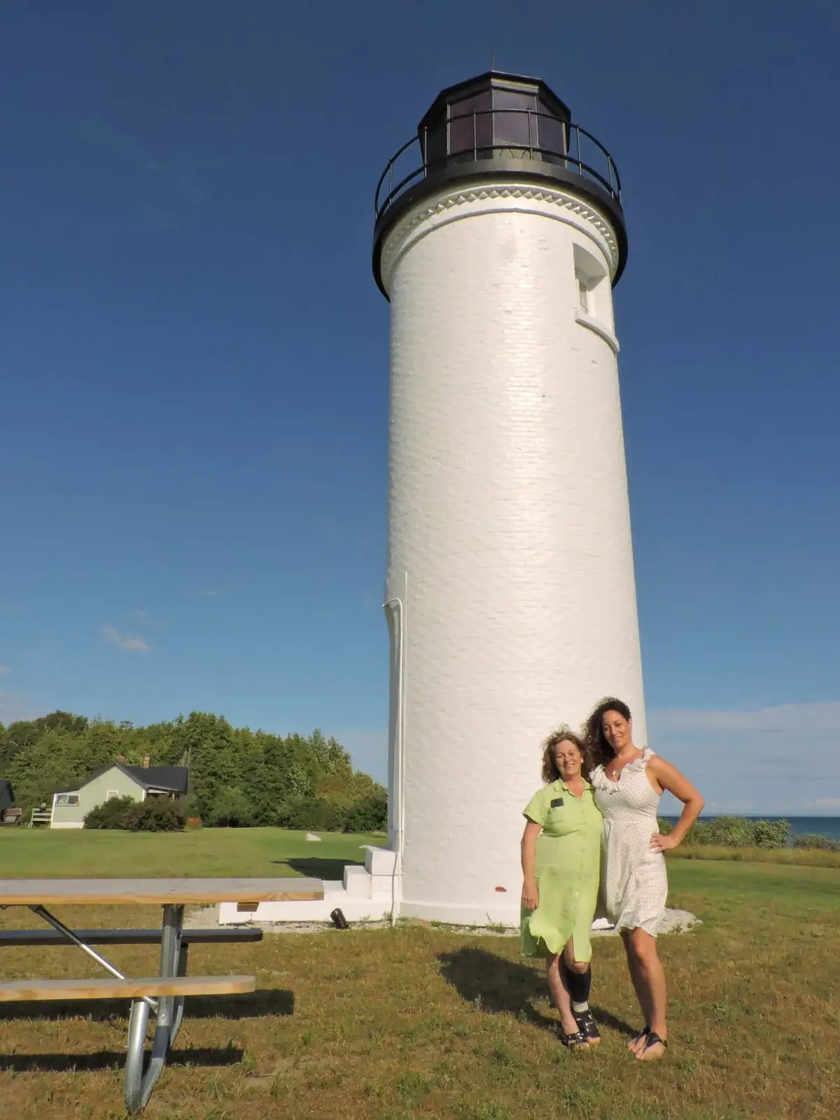 Two women stand beside a tall white lighthouse under a clear blue sky.
