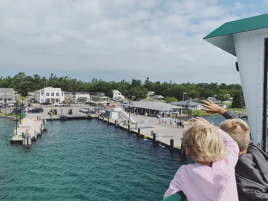 Passengers wave from a vessel toward a waterfront dock lined with buildings, vehicles, and people.
