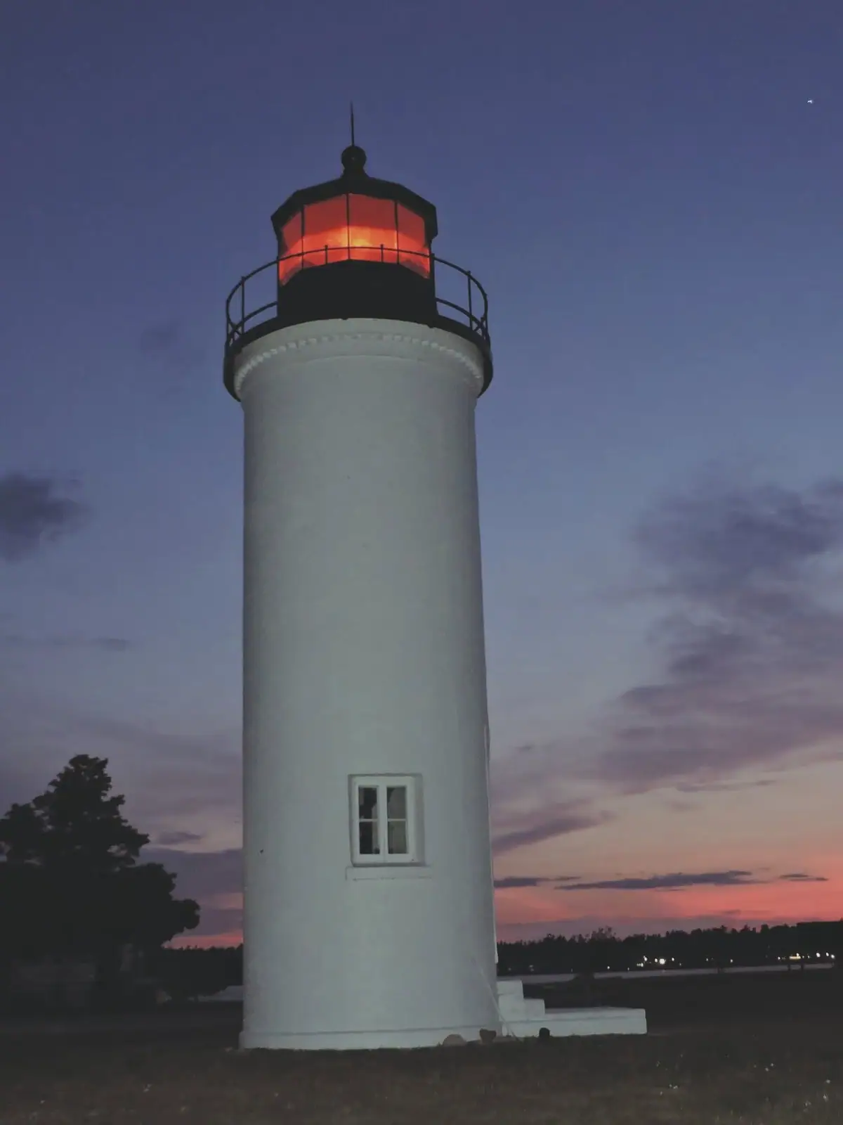 A white cylindrical lighthouse with a glowing red lantern stands against a dusky sky.