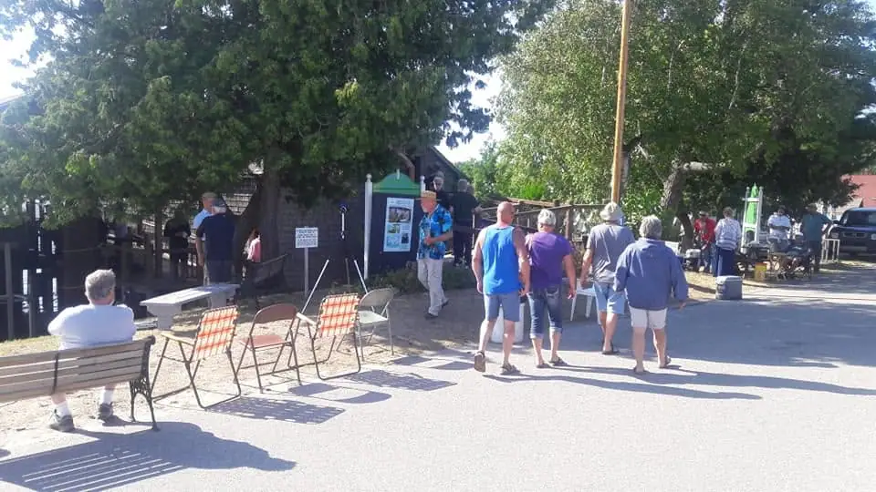 People gather beside folding chairs and an informational display in a sunny, tree-lined paved area.