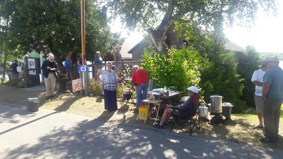 People gather beside a wooden building and a food table beneath large trees, with an OPEN sign visible.