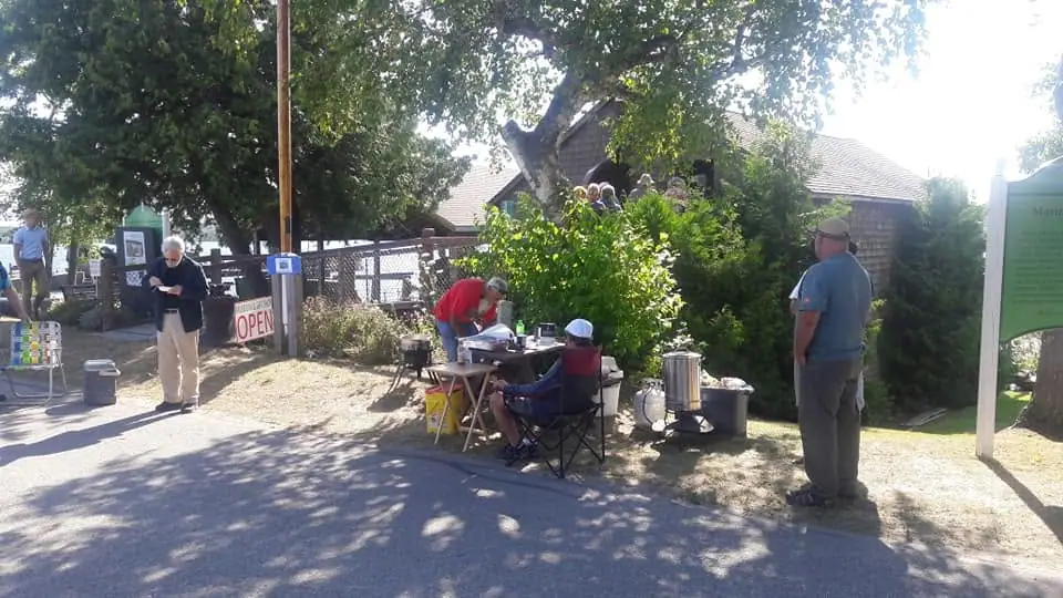 Adults gather beside a building around a folding table with beverage urns and event signs.