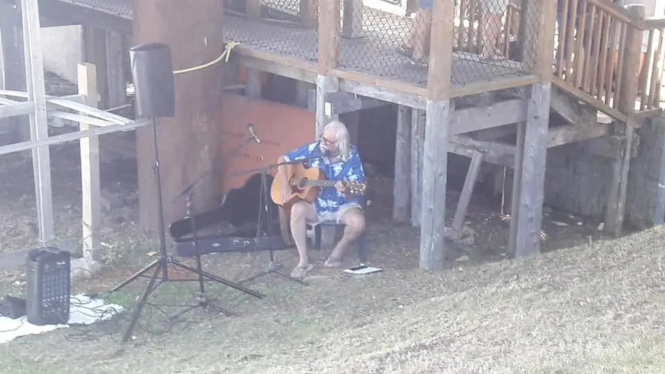 A seated guitarist performs outdoors beneath a raised wooden deck beside a microphone and speakers.