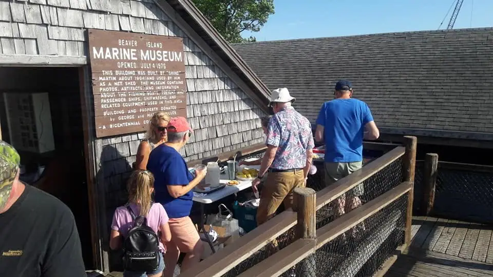 People gather around refreshments on a deck beside a shingled building with a large museum sign.