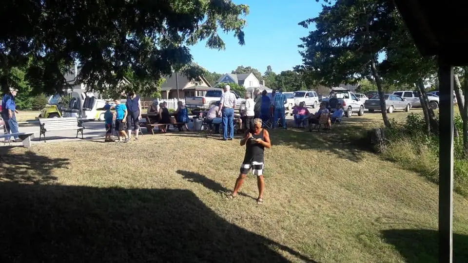 People gather around benches and tables on a grassy lawn while cars and houses stand behind them.
