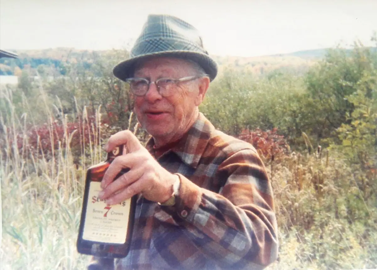 An older man in plaid clothing holds up a bottle while standing among tall grasses and shrubs.