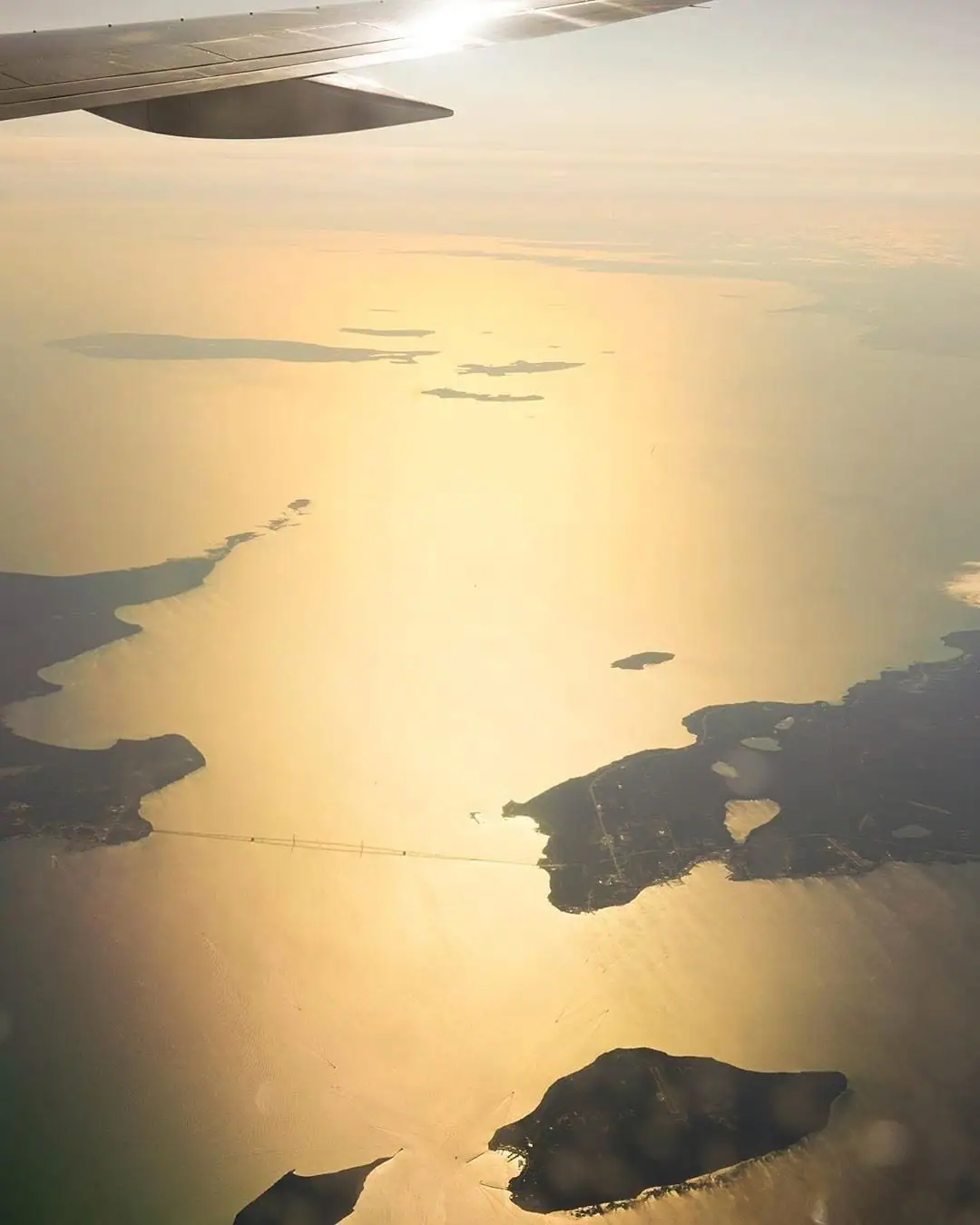 Aerial sunset view of a bridge, several landmasses, and an airplane wing over sunlit water.