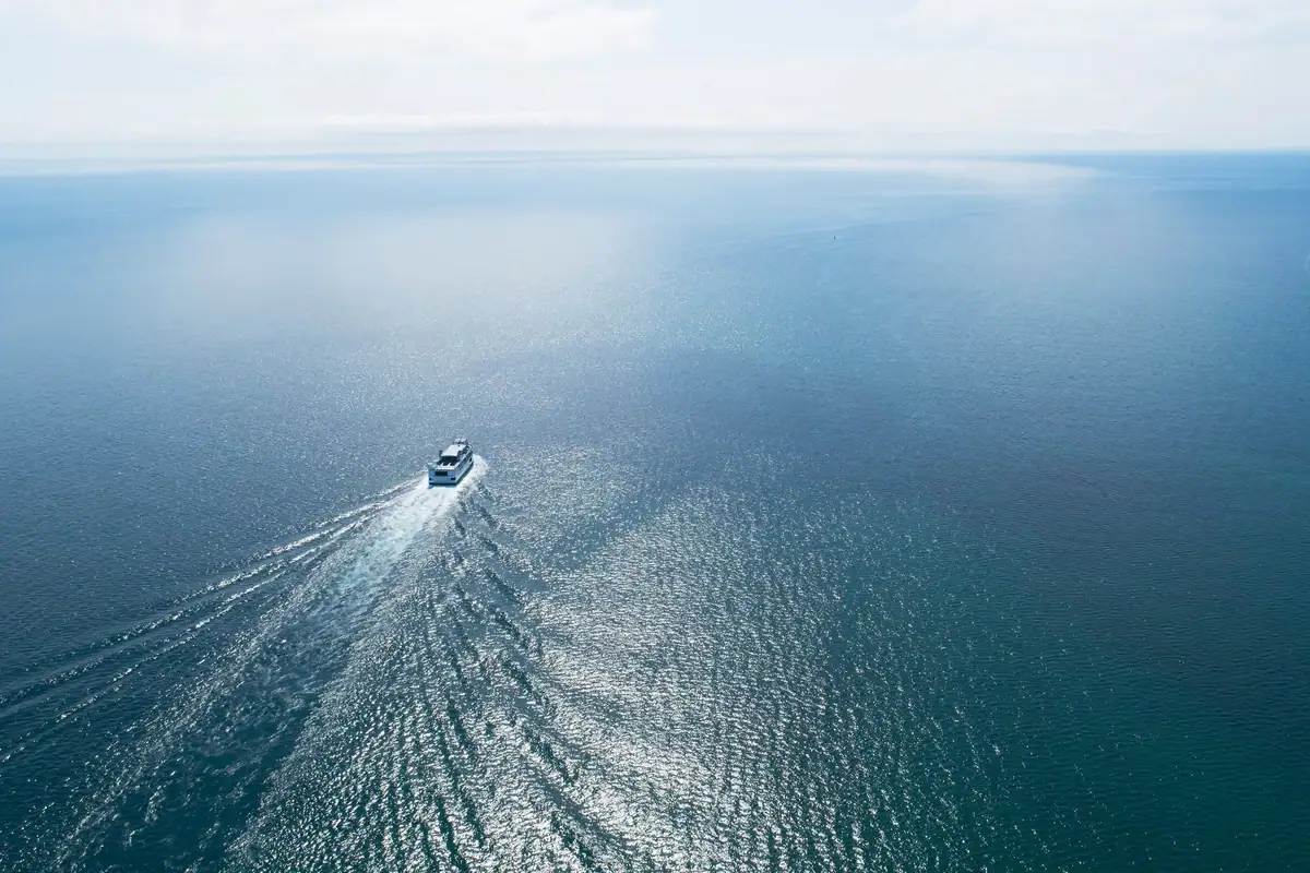 Aerial photograph of a white ferry traveling across open blue water, leaving a broad wake.