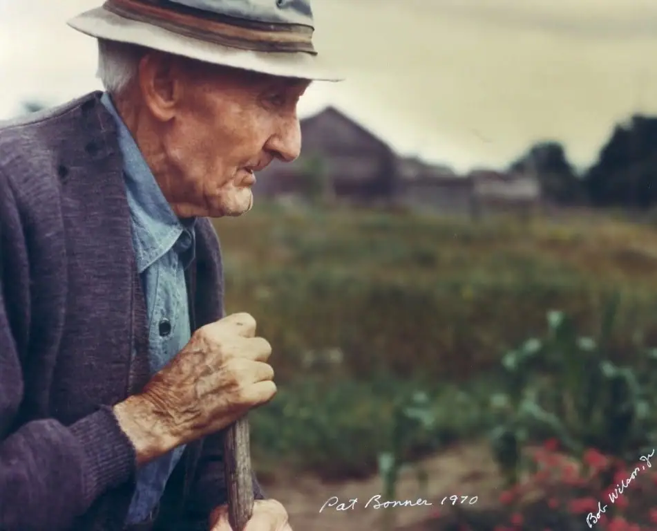 Profile portrait of an elderly man in a hat and cardigan holding a walking stick beside a cultivated field.