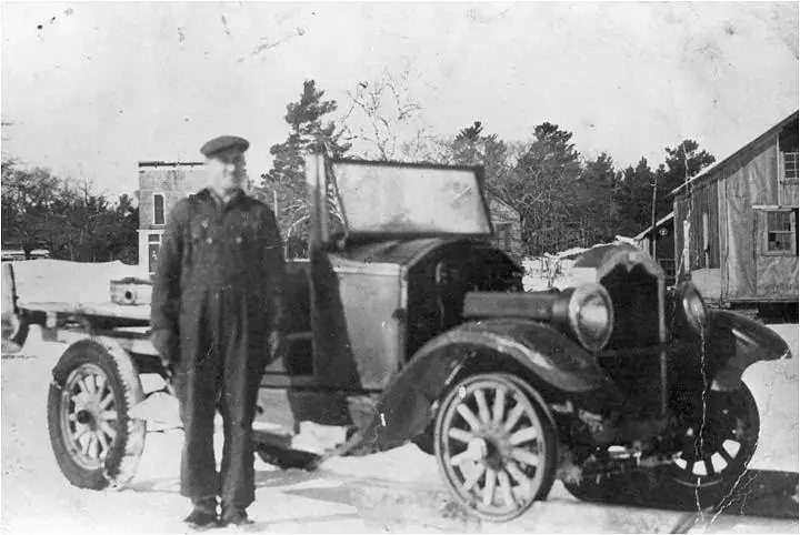 A man stands beside a vintage pickup truck in a snowy yard near wooden buildings.