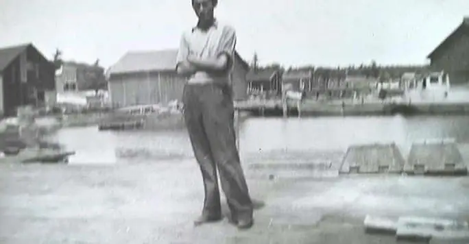 A man stands with folded arms on a waterfront, with docks and buildings behind him.