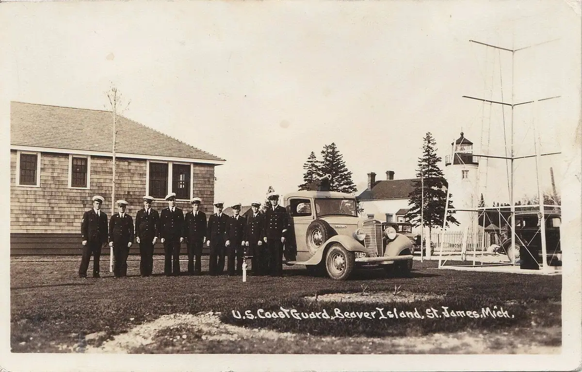 Uniformed men pose beside a vintage automobile, shingled buildings, a lighthouse, and a tall antenna structure.