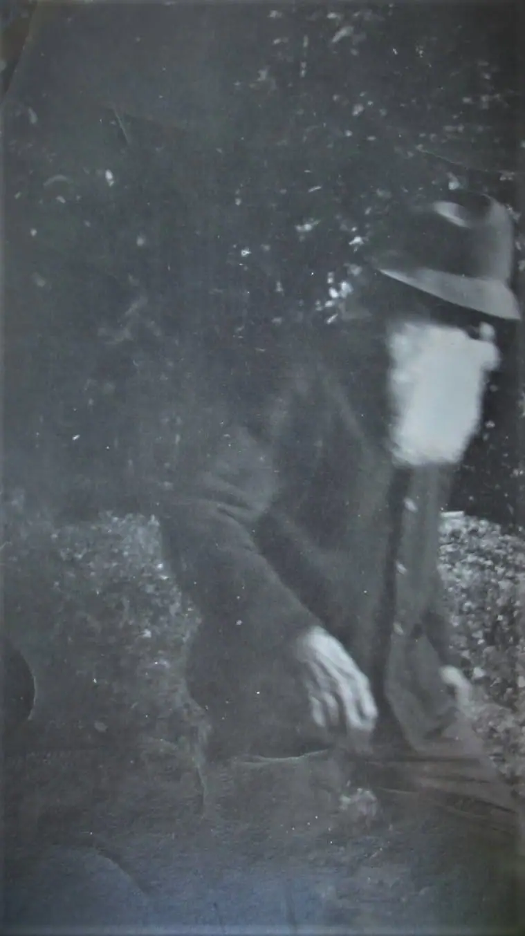 Grainy black-and-white portrait of a bearded person in a broad-brimmed hat and dark coat.