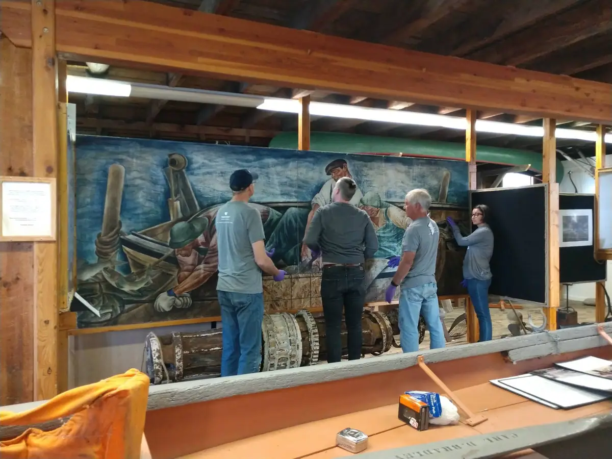 Four people wearing gloves work beside a large painted maritime mural inside a wood-framed building.