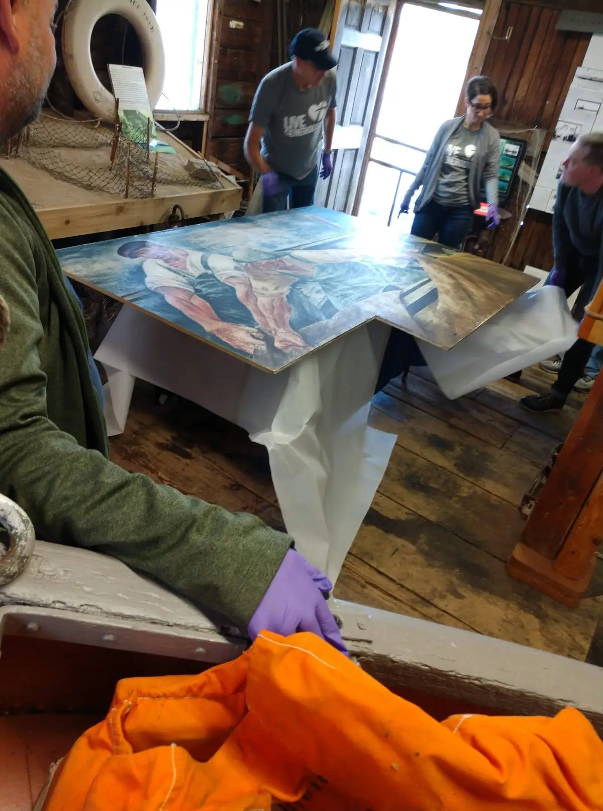 People in gloves handle a large painted panel on a plastic-covered table inside a wood-paneled room.