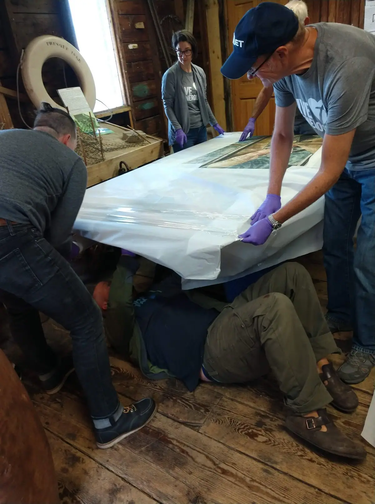 People in purple gloves wrap a large flat framed object inside a wooden room.