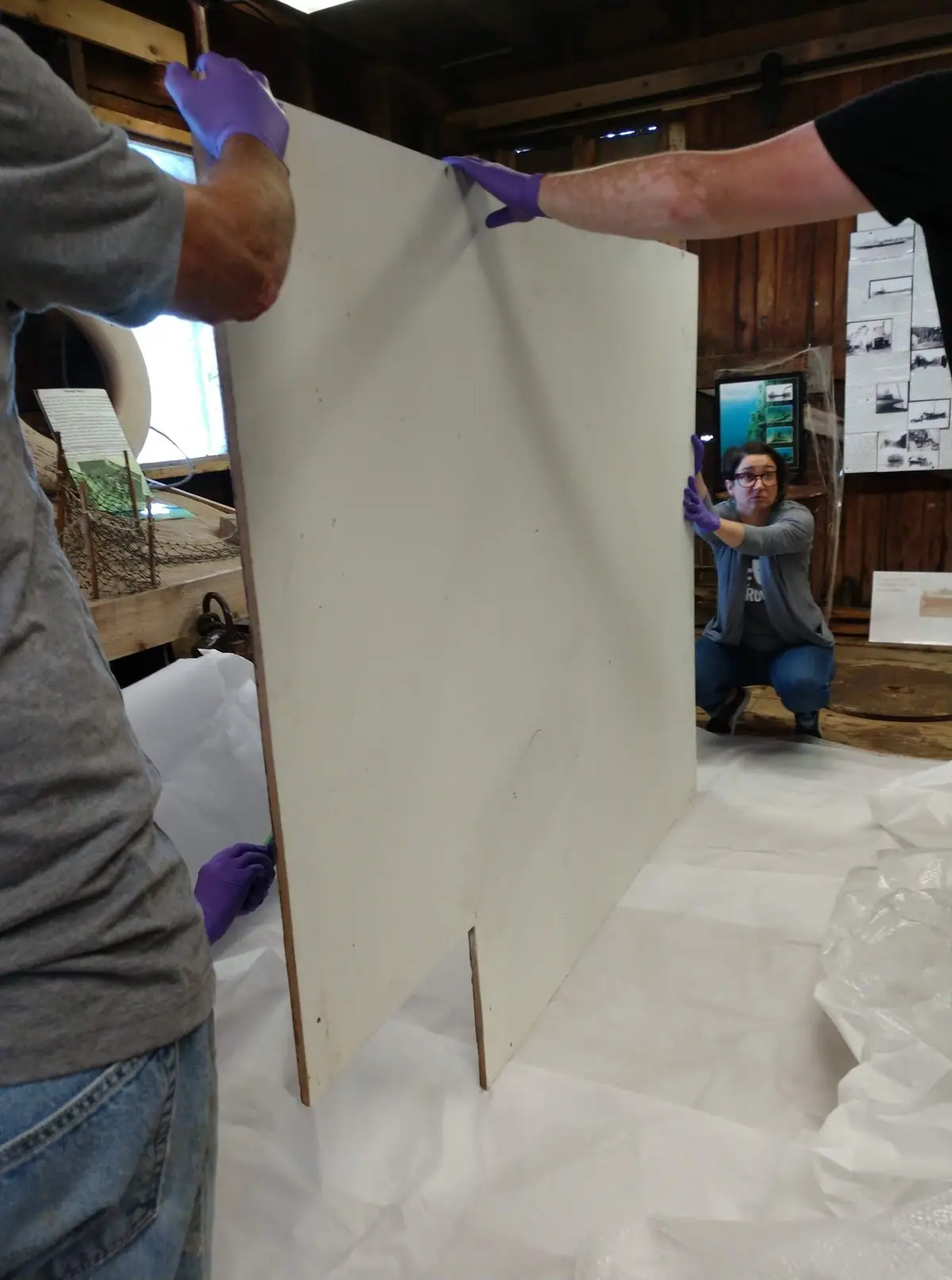 Three people in purple gloves handle a large white panel inside a wood-paneled workspace.