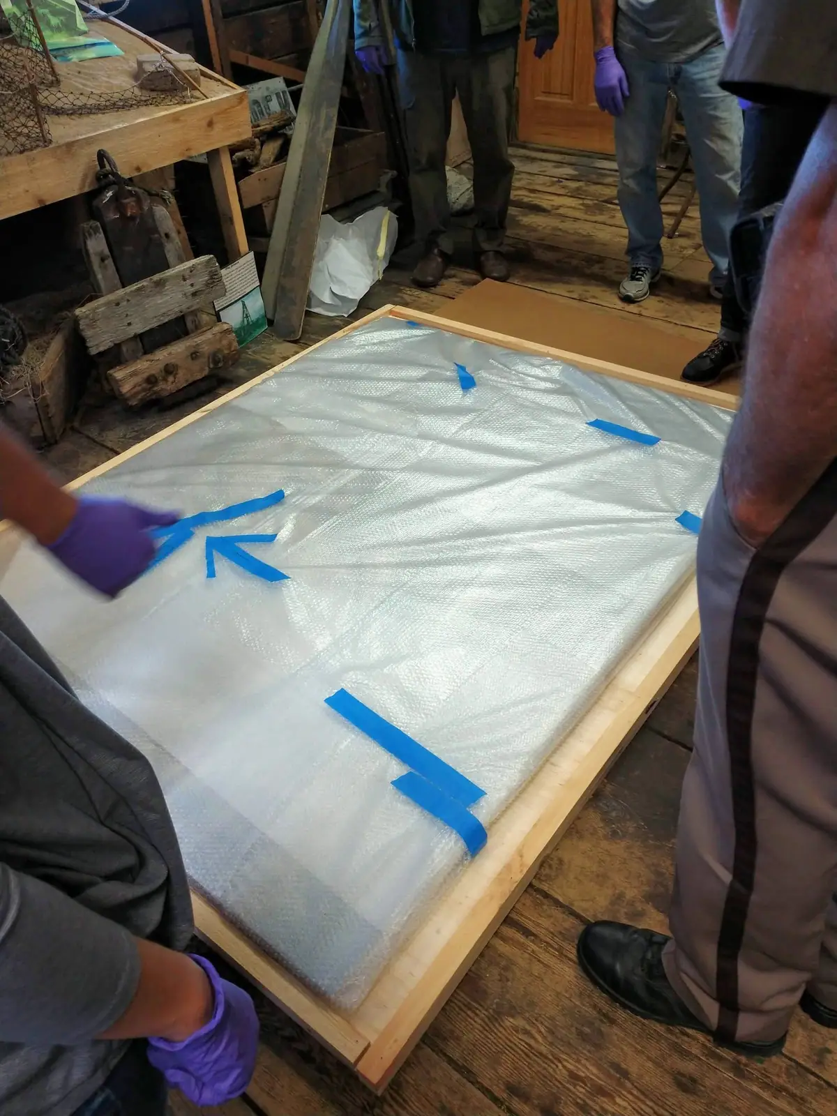 People in a workshop wrap a large rectangular object with translucent material and blue tape.