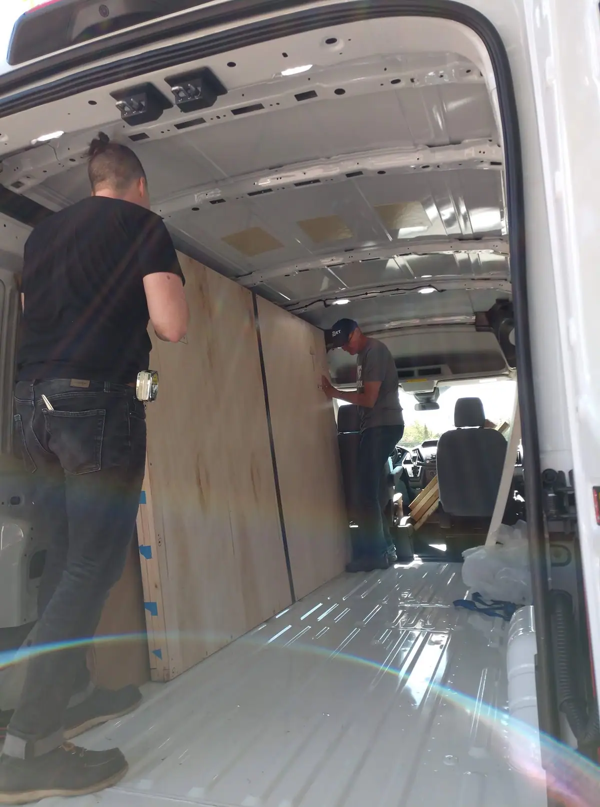 Two men work inside a white cargo van, fitting large wooden panels along its interior walls.