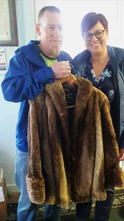 Two people pose indoors while holding a long brown fur coat on a hanger.