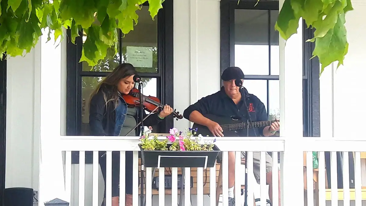 Two musicians perform violin and guitar on a porch with a flower planter.
