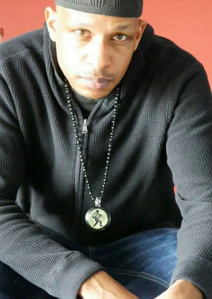 Portrait of a seated man wearing a dark knit cap, textured zip-up sweater, and long beaded necklace.