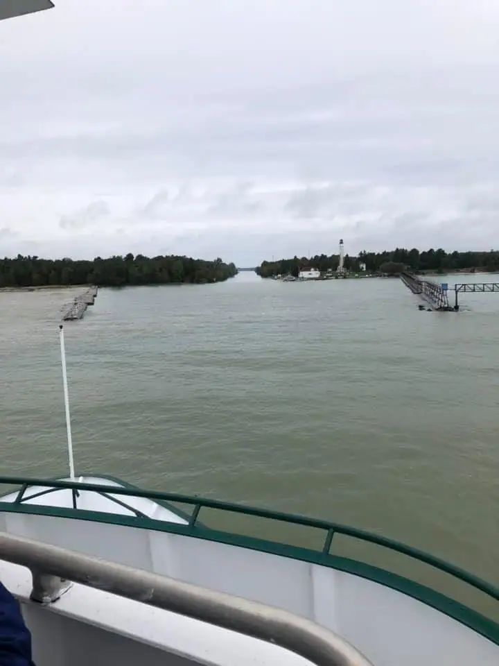 View from a vessel toward a lighthouse, wooded shoreline, and parallel piers beneath an overcast sky.