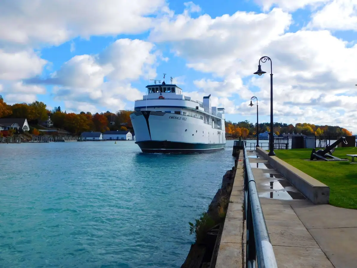 A large white ferry approaches a concrete pier beside autumn-colored trees and ornate dock lamps.