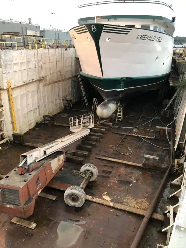 A large white ferry sits in a dry dock while a boom lift and other equipment surround its bow.