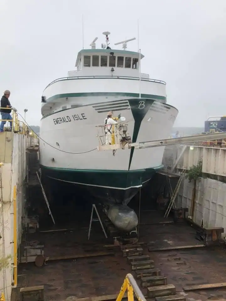 A white passenger ferry is in a dry dock while two workers attend to its hull.