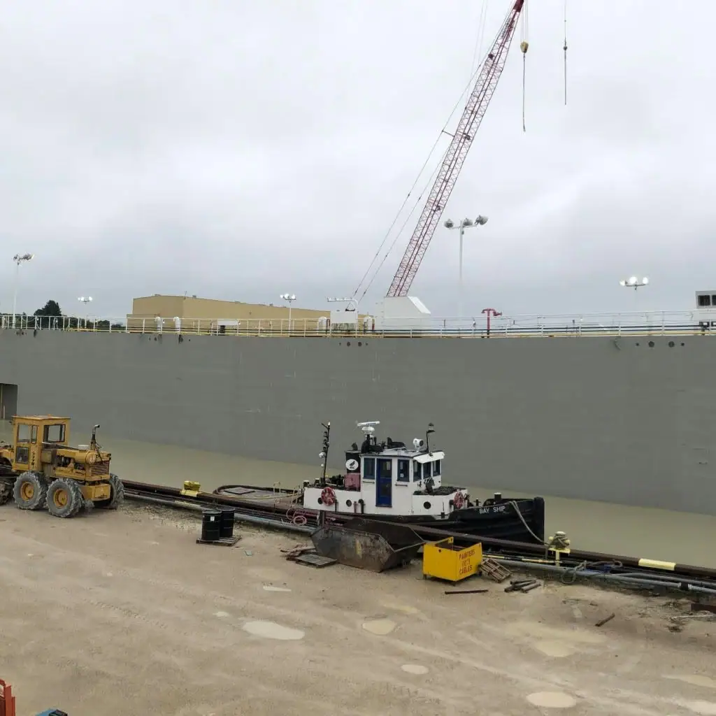 A gray vessel under construction stands behind a small tugboat, crane, and heavy equipment at a muddy worksite.