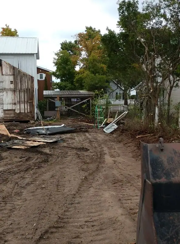 A muddy construction area with a weathered shed, debris, a green ladder, and heavy equipment in the foreground.