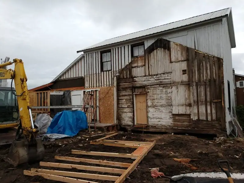 An excavator stands beside a weathered wood-sided building under renovation, with new framing laid out in muddy ground.